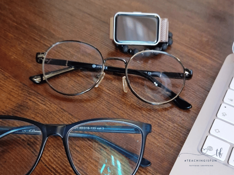 A close-up view of two pairs of eyeglasses, a smartwatch, and the corner of a white keyboard on a wooden desk.